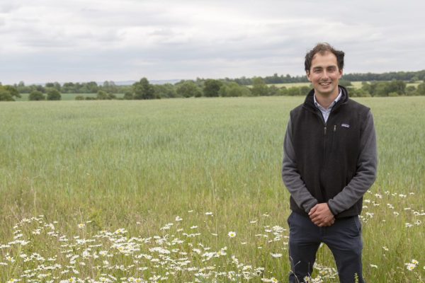 Callum Weir standing in a field margin of oxeye daisies bordering a field of wheat